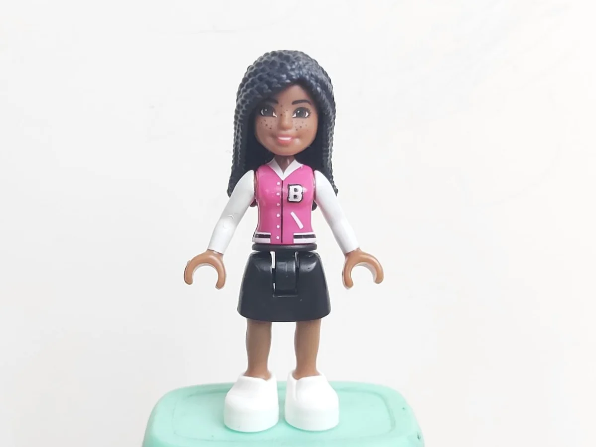 Barbie Micro-Doll, medium brown skin, black afro-textured hair, barbie pink jacket with white arms and B logo, black simple skirt, white shoes, dark brown eyes, freckles