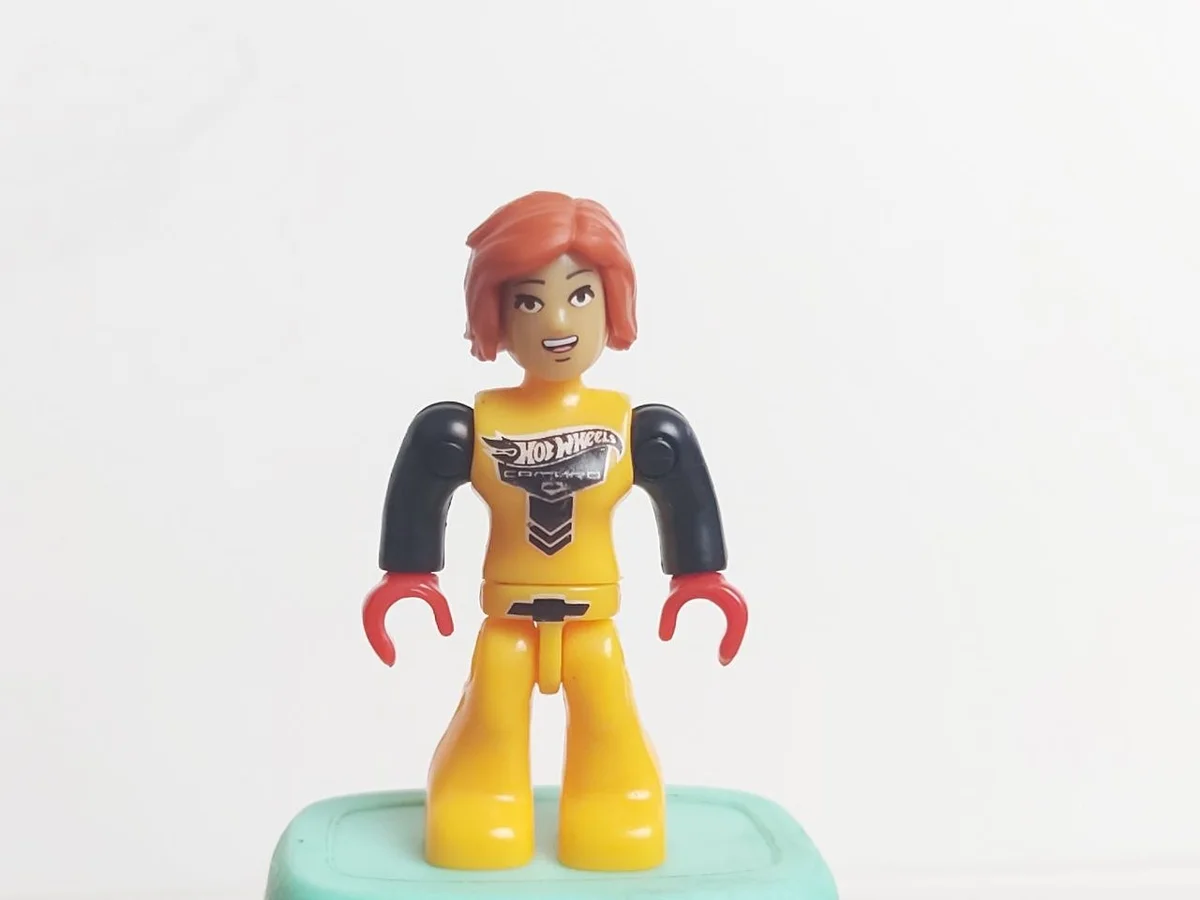 Driver Female, dark orange hair, black arms, red hands, light orange jumpsuit with black and silver "HOT WHEELS CAMARO" logo in Yellow, Orange